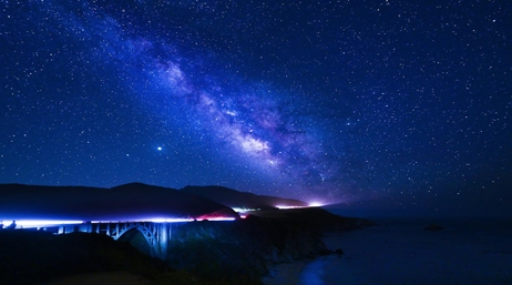 A view of a bridge on the coast at night, illuminated by streak lights from cars, and under a starry blue and purple sky