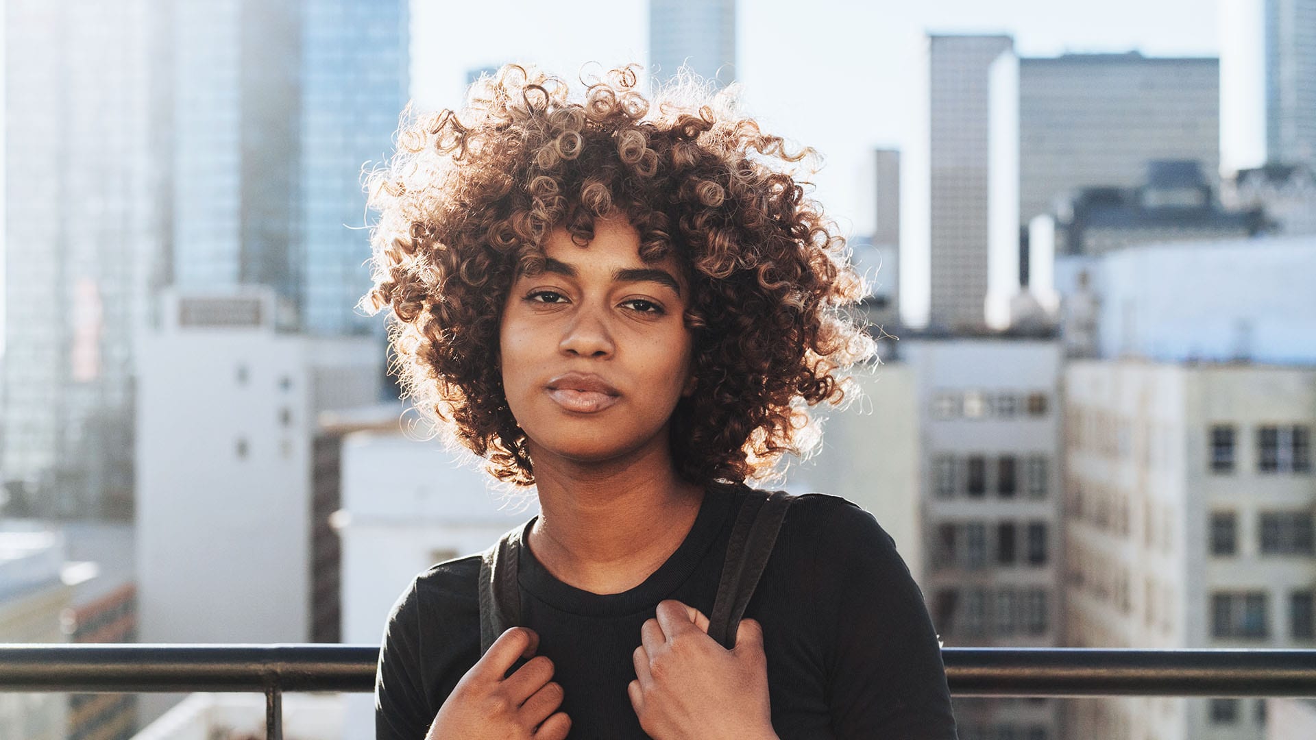 A person of color standing in the sun with a cityscape in soft focus in the background 