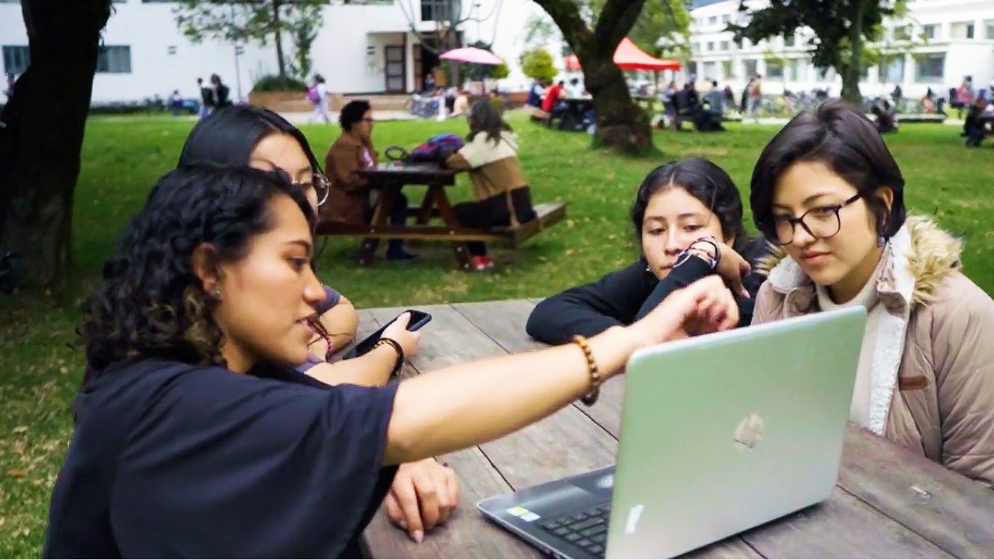 A group of students discussing a shared laptop display on a picnic table in a large green park