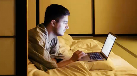 Uma pessoa vestida casualmente, deitada de bruços na cama, usando um laptop em um quarto bem iluminado, com paredes cor de mel e painéis pretos