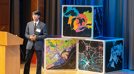A student in a gray suit speaking onstage at a conference with large blocky map-themed decor