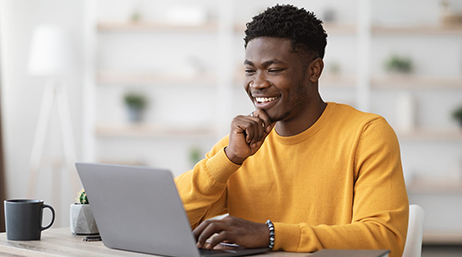 A person wearing a yellow long-sleeved tee shirt smiles as they read an open laptop display in a sunlit home office