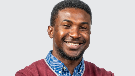 Portrait of Chima Okoli smiling in a blue collared shirt and red sweater with a pale gray background