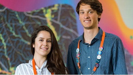Two conference attendees with their lanyards and pins on smiling in front of a backdrop