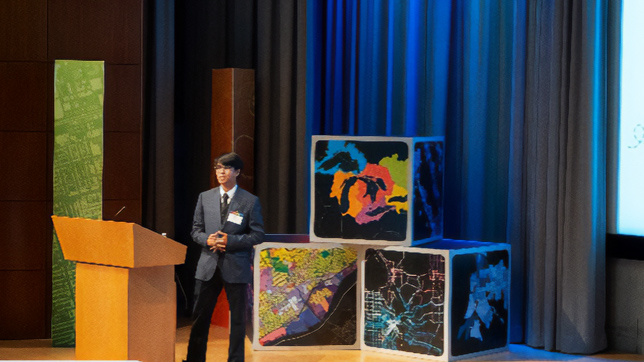 Young professional wearing a gray and black suit on stage at the Esri campus auditorium giving a presentation
