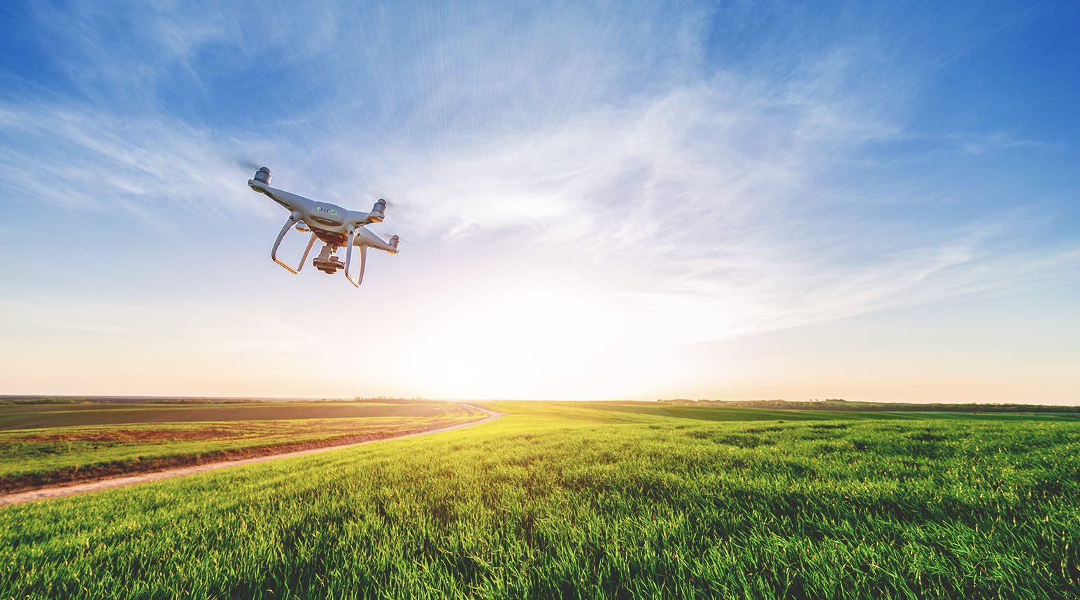 A white drone flies in a blue cloud-streaked sky over a green grassy field