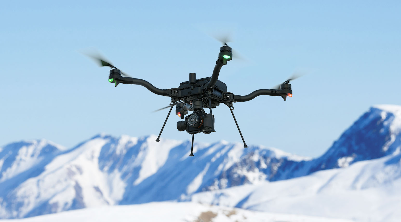 A white drone flies in a clear blue sky over snowy mountains