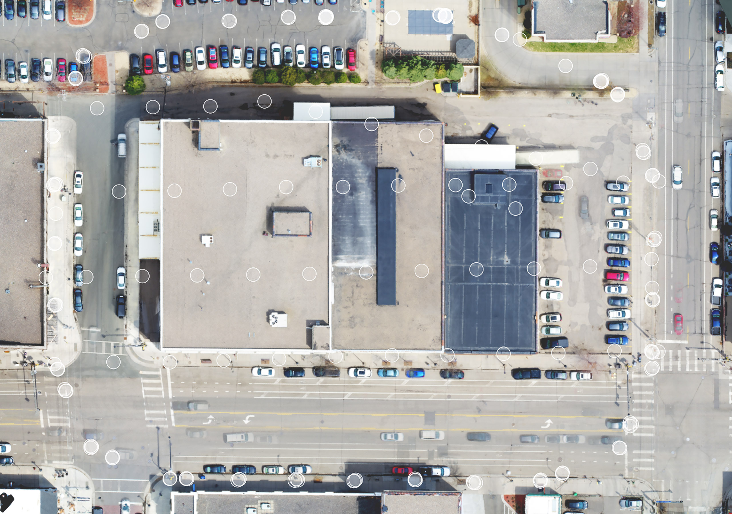 A nadir image of a commercial building on the corner of a city intersection and cars parked in two parking lots