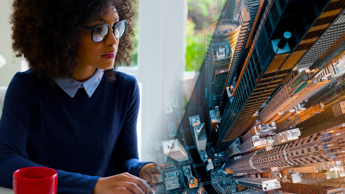 A composite image with a person in a blue sweater using a computer in a bright office alongside an aerial photo of towering modern skyscrapers