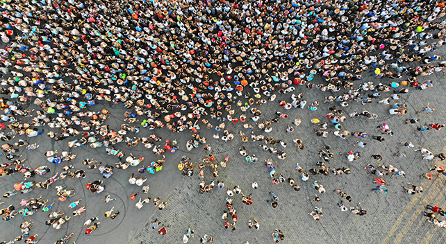 An aerial photo of a colorful crowd of people tightly grouped on a gray concrete and cobblestone road
