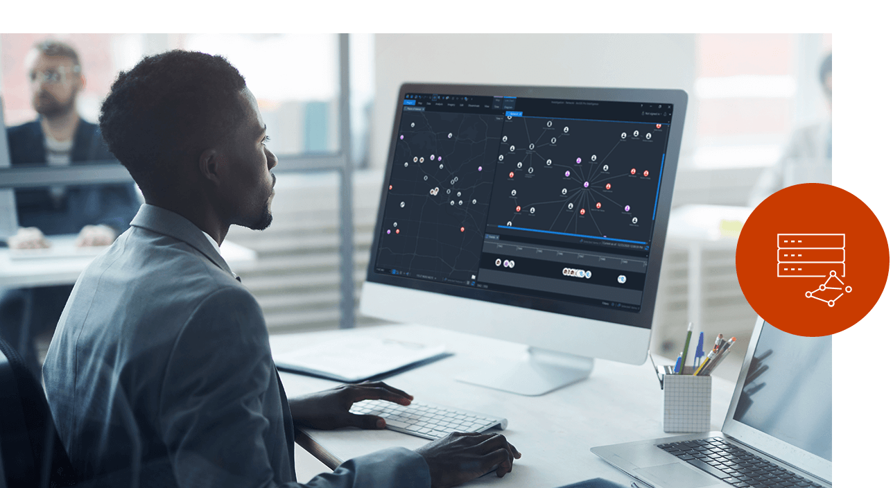 Jovem trabalhando em um computador com um grande monitor mostrando dados simbolizados em um mapa e em um gráfico de links 