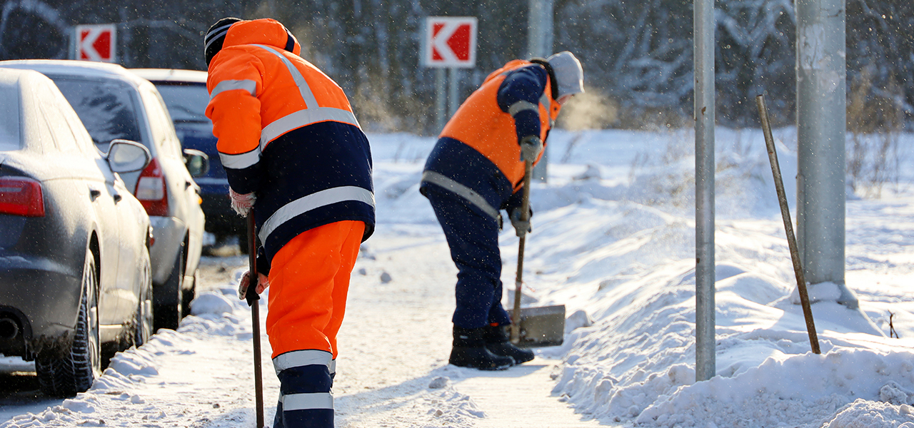 Two people wearing heavy winter clothing and orange safety gear shoveling deep snow along a busy road