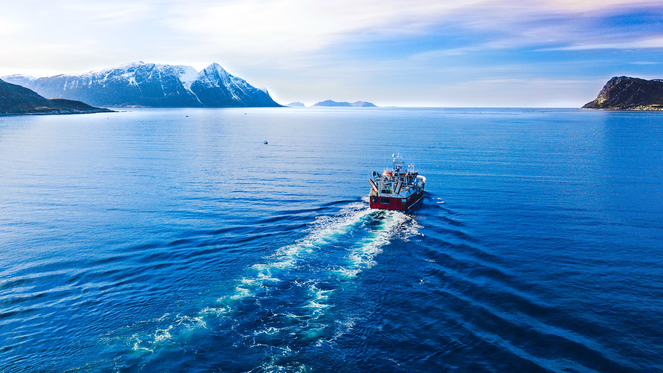 A small research vessel traveling across deep blue water, leaving a wake behind as it heads toward distant coastal mountains