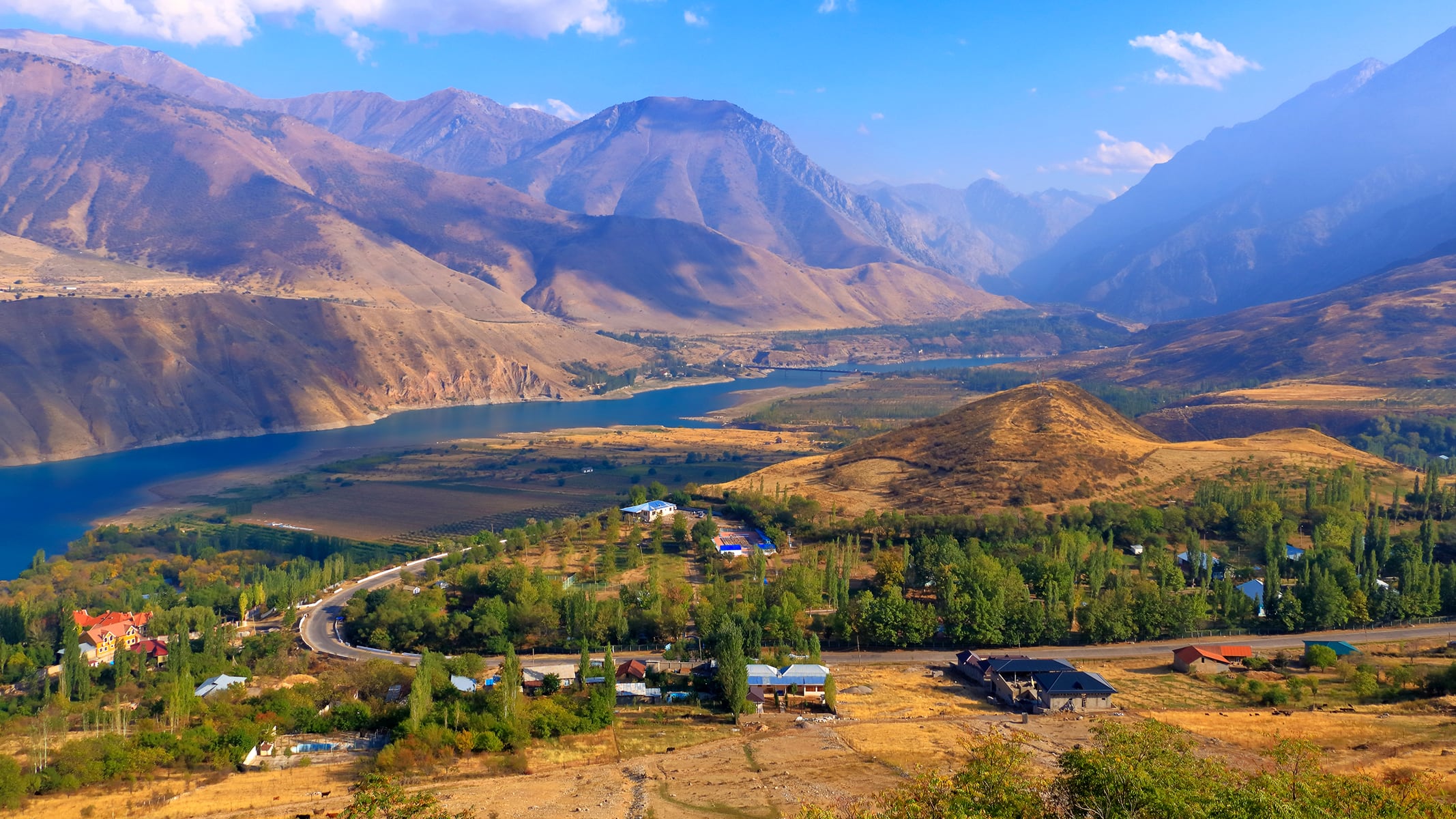 A wide valley with agricultural fields, a river, and tall mountains rising in the background under a partly cloudy sky