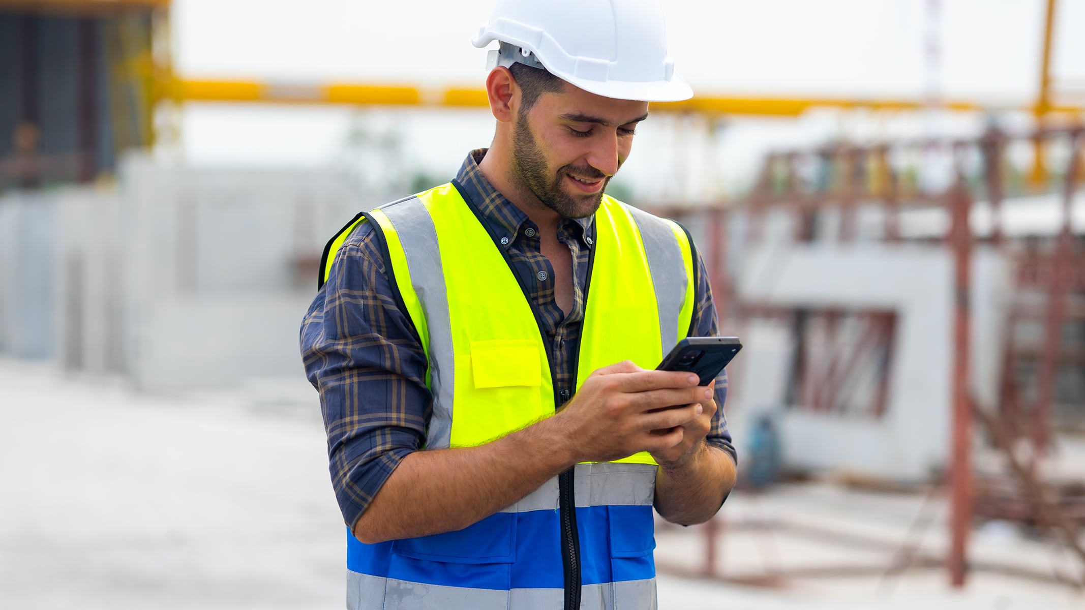 A man wearing a safety vest is focused on using his smartphone outdoors.