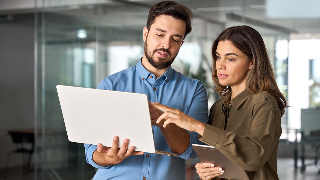Two professionals standing in a modern office, collaborating while looking at a laptop and holding a tablet, with a play button indicating the image can be clicked to start a video