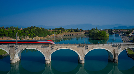 A red and silver commuter train speeding along a bridge with a row of arches over still blue water beneath a clear blue sky