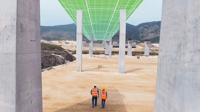 Two people in orange safety vests and hard hats in a bare brown field with mountains in the distance, walking under an overpass overlaid with a graphic of a green and white grid