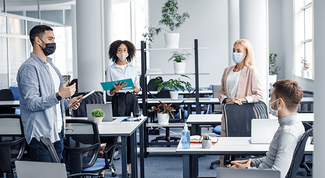 Quattro lavoratori socialmente distanti in un ufficio che indossano maschere protettive, che hanno una conversazione