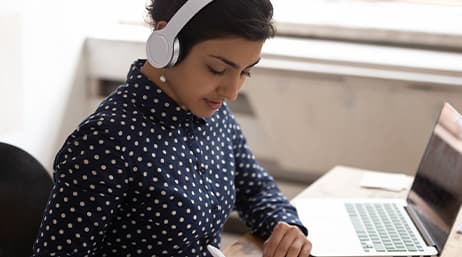 A person with wireless headphones on, writing notes with a computer next to them