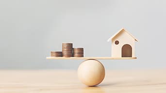 A wooden plank carrying coins and a model of a house, balancing on a ball.