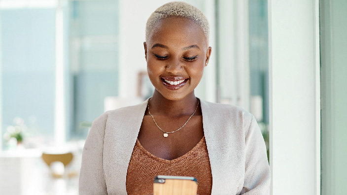 Una persona sorridente che indossa una giacca chiara sorride guardando un telefono cellulare in mano con un elegante ufficio moderno in vetro