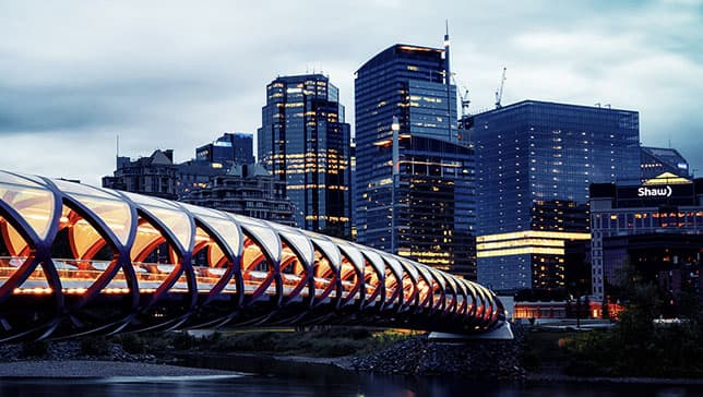 A modern bridge arching over a river toward a sleek city filled with shining skyscrapers brightly lit against a pale evening sky
