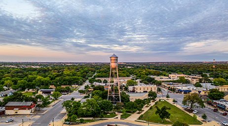 Aerial image of a water tower in the center of a neat sprawling town filled with green trees and small brown buildings under a cloud-streaked sunset sky