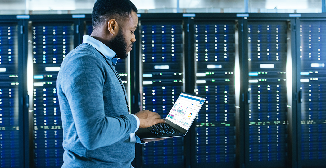 A person standing and using a laptop in front of a wall of server racks overlaid with a play button