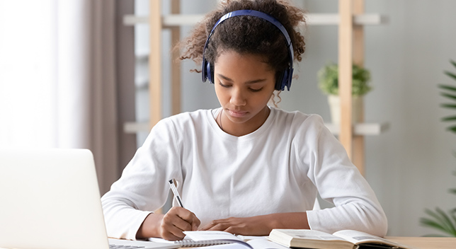 O fetiță cu căști pe urechi, care stă în fața unui laptop și scrie într-un caiet