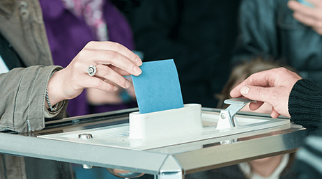 Primer plano de la mano de una mujer vestida con abrigo depositando una papeleta azul en una urna. 