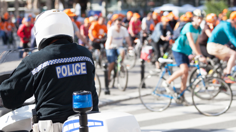 A large group of cyclists wearing orange helmets on a sunlit city street with a police officer on a motorcycle in the foreground