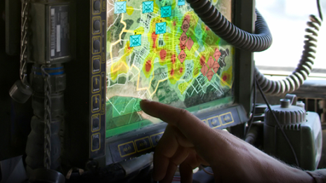 A closeup of a hand pointing to a computer screen surrounded by communications equipment and displaying a colorful map
