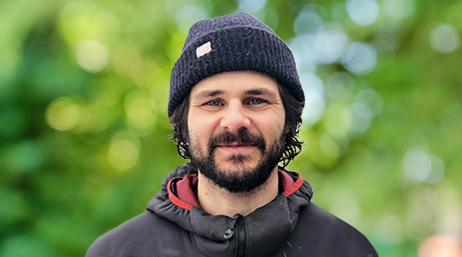 Portrait of Charlie Mix smiling in a black anorak and beanie with sun-dappled trees in the background