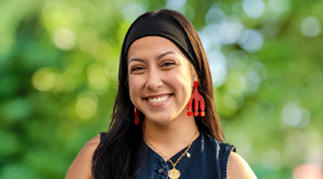 Portrait of Karina Amalbert smiling in a dark sleeveless shirt with sun-dappled trees in the background