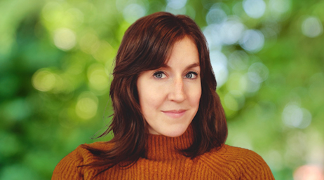Portrait of Lauren Tierney smiling in an orange turtleneck shirt with sun-dappled trees in the background