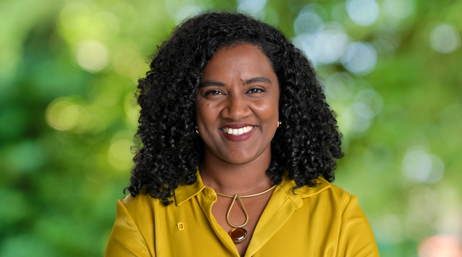 Portrait of Véronique Couttee smiling in a yellow collared shirt with sun-dappled trees in the background