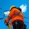A person in orange safety gear and a yellow hard had climbing a ladder to reach power lines at the top transformer with a bright blue sky overhead