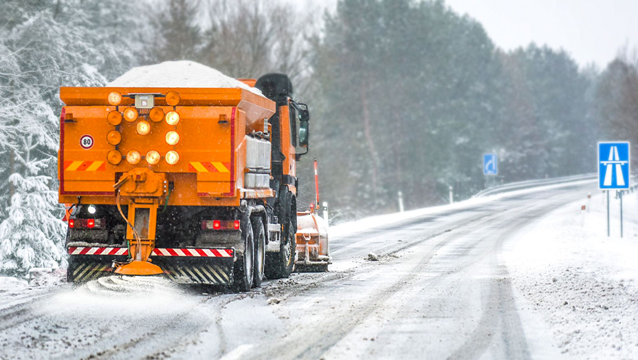 An orange snowplow driving down a snowy tree-lined road