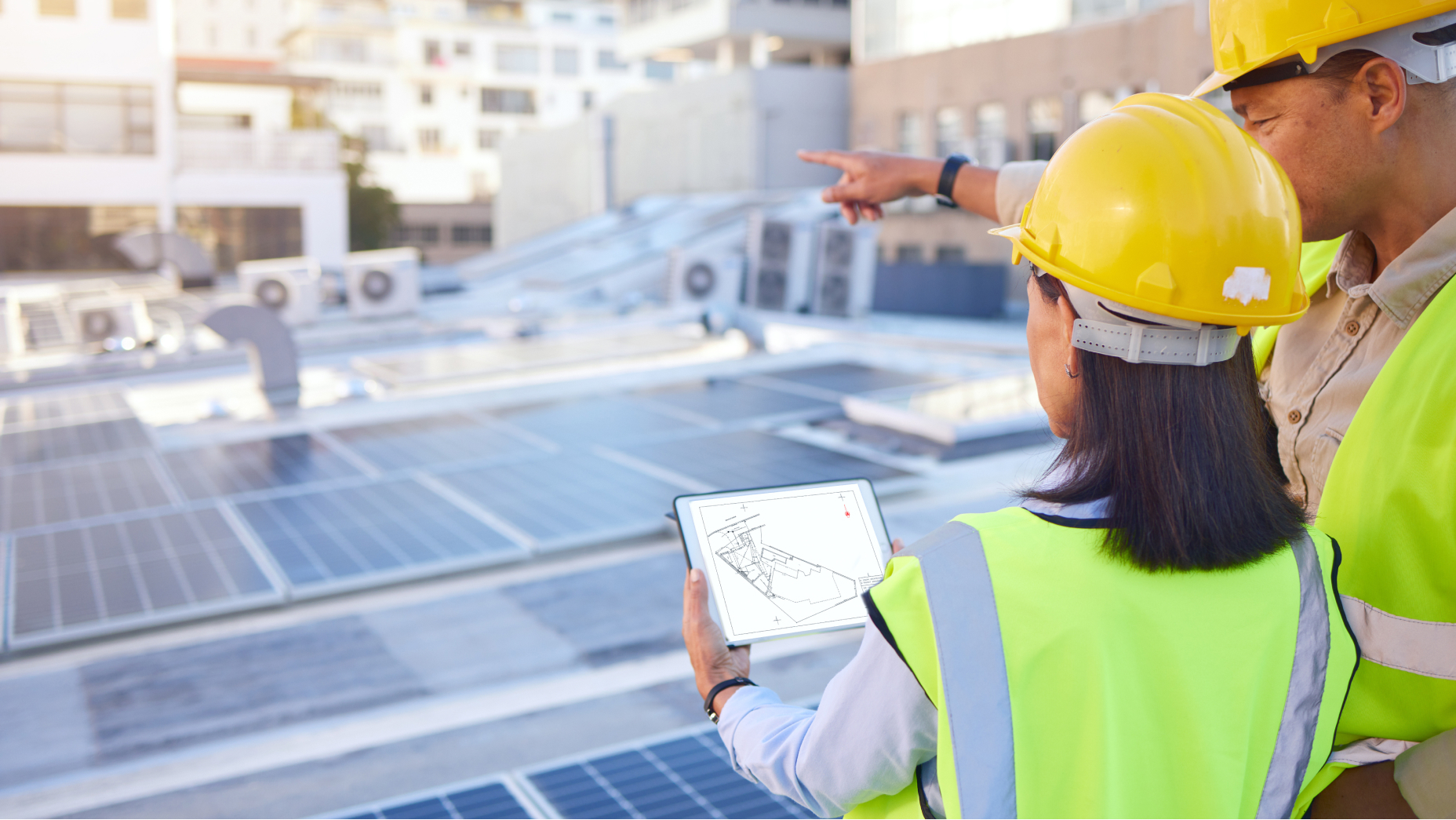 Ein Mann und eine Frau mit Schutzhelmen und gelben Westen mit einem Tablet vor großen Solarpaneelen 