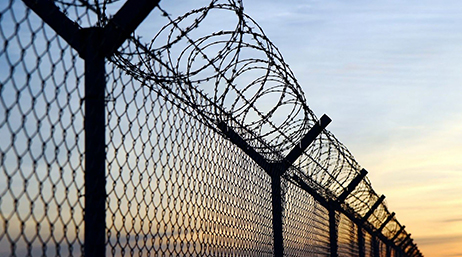 A chain link fence topped with barbed wire stretches into the distance, silhouetted against a pale blue and orange sunset