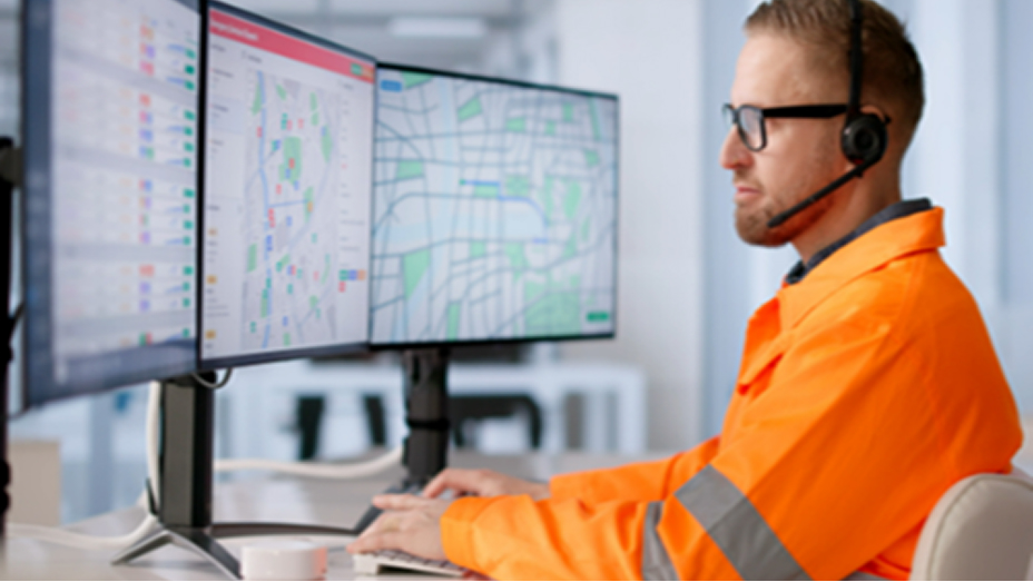 A person wearing an orange visibility jacket and headset sitting at a desk looking at multiple monitors displaying maps and data
