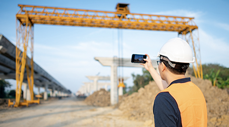 A mobile worker wearing a white hard hat and an orange vest taking a picture of a construction site