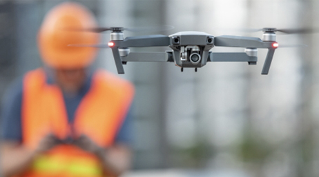 A drone flying in midair in sharp focus with a mobile worker wearing an orange hard hat and vest in the background