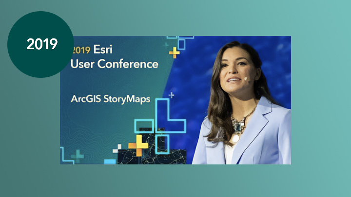 Eine Person in einem blassblauen Anzug mit Headset, die auf einer Bühne vor einem tiefblauen, mit geometrischen Formen in Grün und Gold sowie dem Text "2019 Esri User Conference – ArcGIS StoryMaps” überlagerten Hintergrund spricht