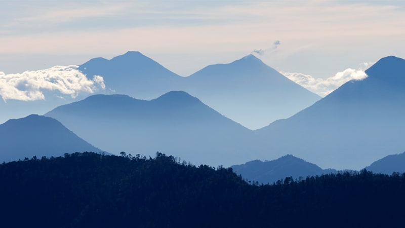 A mountainous landscape shot with dark blue mountains in the forefront, lighter blue mountains in the middle, and snow-capped mountains  under a cloudy sky in the background