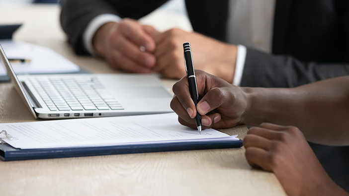 Duas pessoas assinando um contrato em uma mesa ao lado de um laptop aberto