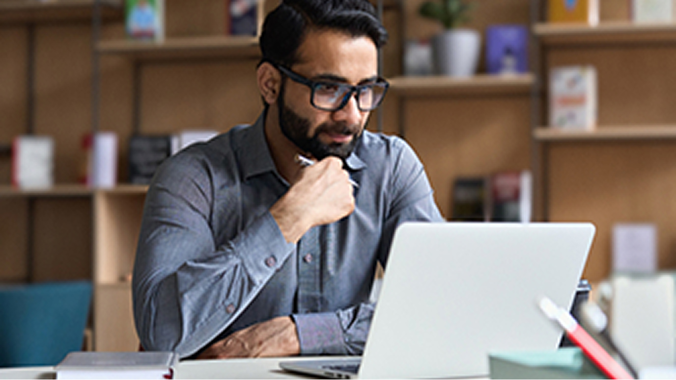 A man wearing glasses sits at a desk, concentrating on his work at a laptop computer. 