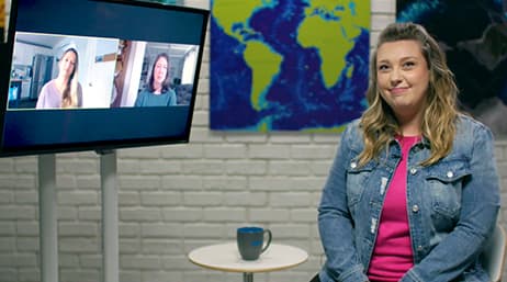 A person sitting and smiling next to a raised monitor that displays two people in a video chat 