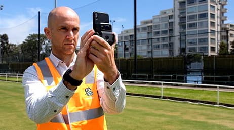 Un lavoratore che indossa un giubbotto di sicurezza che guarda il suo telefono su un manto erboso, sovrapposto da un telefono touchscreen che mostra la soluzione di misurazione Spile in ArcGIS Survey 123